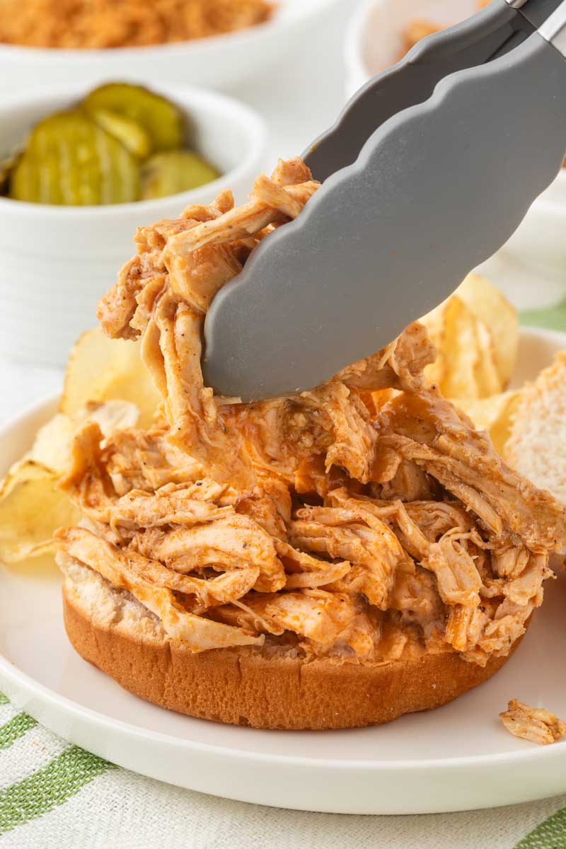 Tongs grabbing slow cooked spicy chicken and placing it on a bun to make sandwiches