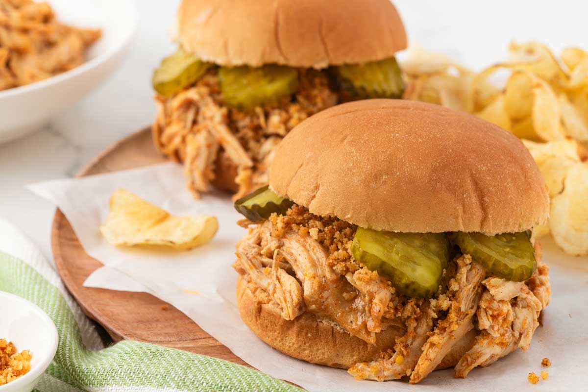 slow cooker nashville chicken on a bun, surrounded by chips and pickles peeking out